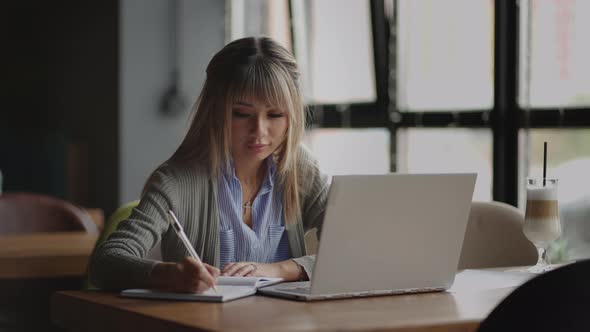 A Young Asian Woman Writes with a Pen Looking at the Laptop Screen Sitting at the Table