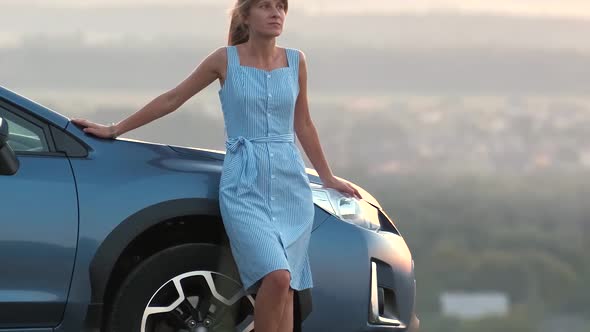 Happy young woman driver in blue dress enjoying warm summer evening standing beside her car.