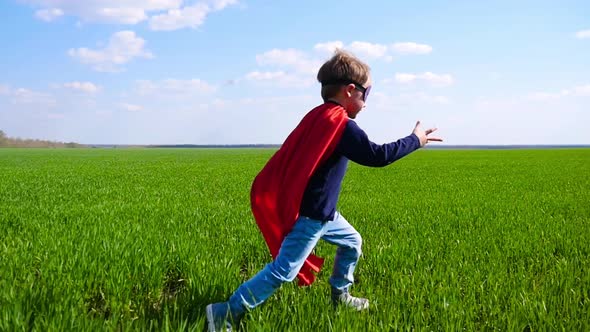 A Little Happy Child in a Superhero Costume, a Red Cloak and a Mask Runs on Green Grass Against a