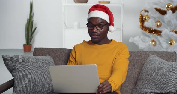 Afro American Man Black Man Wears Santa's Hat Sits with Laptop on Sofa at Home Near Cristmas Tree