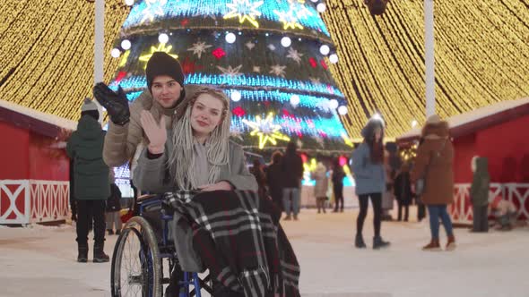 Smiling Young Woman in a Wheelchair and Her Boyfriend at Christmas Party on the Streets Waving Hands