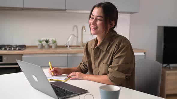 Female Student Watching a Lecture Webinar Using a Laptop Sits at the Table and Writes with a Pen