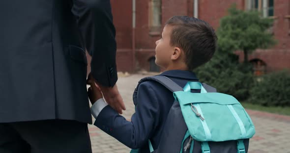 Backside View of Little Kid with Backpack Holding Hand in Hand His Father in Suit. Male Person