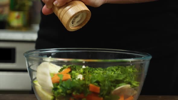 Closeup of a Woman's Hand Begging Black Pepper From a Wooden Pepperpot Into a Salad in a Glass Bowl