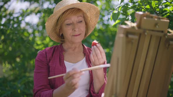 Portrait of Positive Talented Senior Woman Painting Strawberry in Slow Motion Standing at Easel in