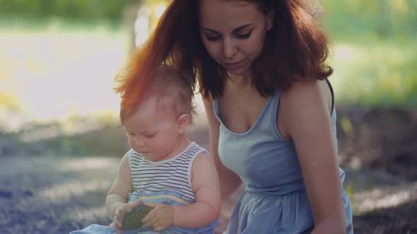 mother and baby sit in a park or in a forest on the ground looking at twigs and cones