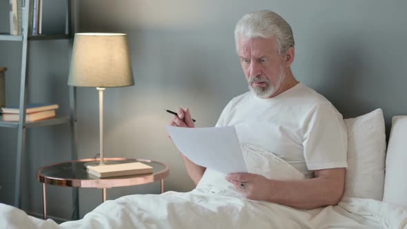 Old Man Reacting to Loss on Documents in Bed