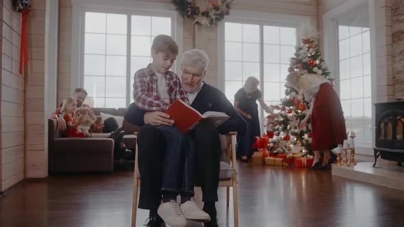 Grandad Reading a Book with Grandson Sitting on a Chair at Christmas Celebration