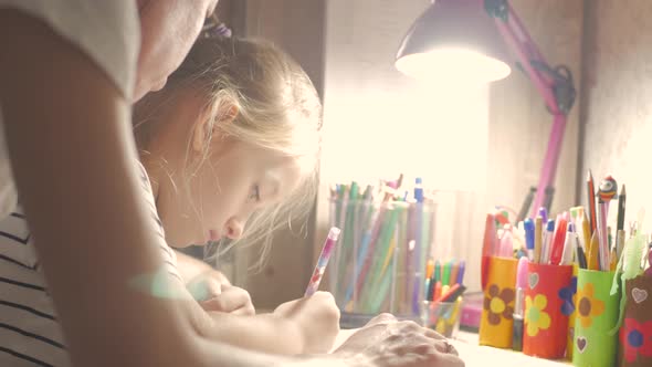 Young Mother Helps Her Daughter with Her Homework at on the Table Under the Light of a Lamp.