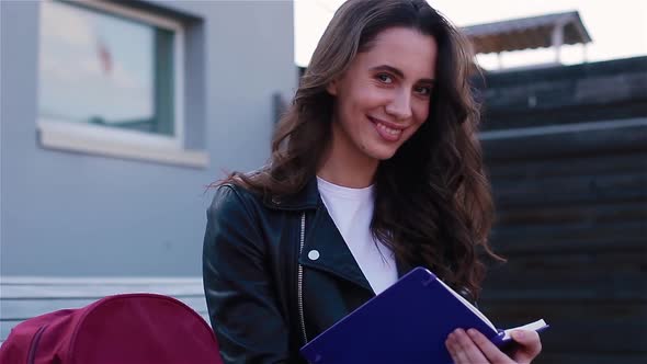 Female Student Writing in Notebook Sitting Outside on the Modern Wooden Bench