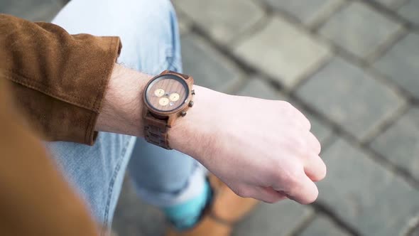 Male wrist with a wooden fashionable watch and brown jacket,jeans,city.