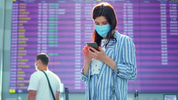 Female Air Passenger in Mask, Standing Against Departure Board at Airport. She Is Checking Flight