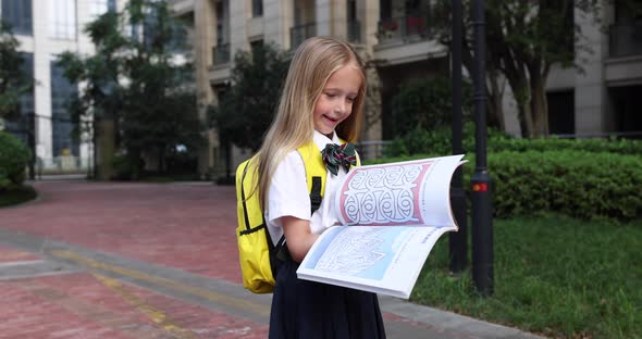 Happy Little Caucasian Blonde Girl Seven Years Old in Uniform with Yellow Backpack Reading Book Near