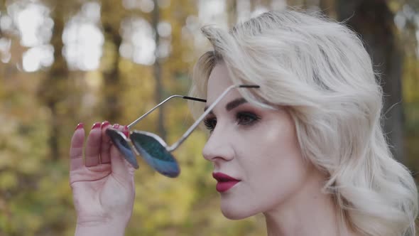 Seductive Blonde with Red Lips Puts on Sunglasses and Looks Straight in Park