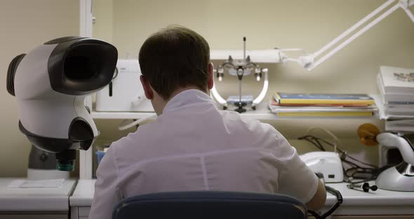 A male dentist sits with his back and looks into a microscope.