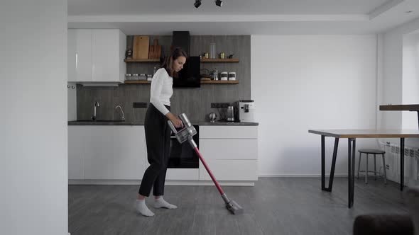Slender Brunette Vacuums in the Kitchen. A Modern Wireless Vacuum Cleaner.
