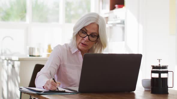Senior woman wearing glasses taking notes and using laptop while working from home