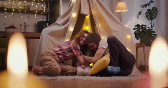 Happy Young Family Sitting on Floor with Teepee and Embracing