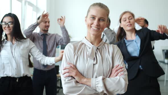 Joyful Blond Lady Smiling at Camera Standing in Workplace Where Businesspeople Dancing Having Fun