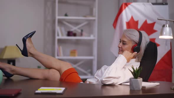 Relaxed Middle Aged Businesswoman Sitting in Office with Feet on Table Listening to Music in