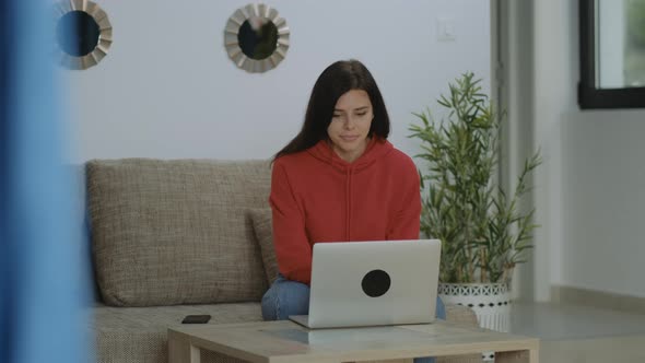 Young freelancer brunette woman in red hoodie open laptop and working, sitting in sofa at home