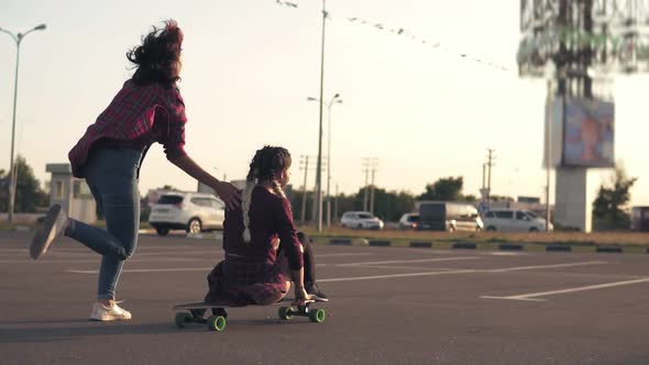 Back View of a Woman Sitting on a Longboard While Her Friend is Pushing Her Behind and Running