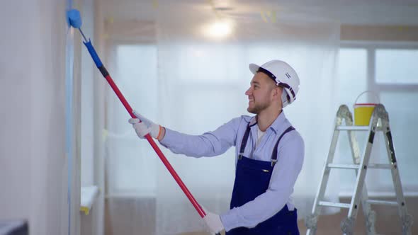 Male Painter in Uniform and Helmet Makes Repairs Indoors and Paints Walls Using Construction Roller