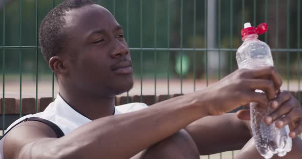 African American Guy Drinking Water, Sitting at Basketball Sportsground