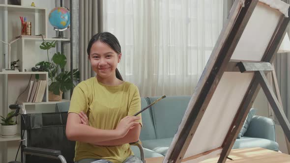 Kid Girl In Wheelchair Holding Paintbrush And Cross His Arms And Smile While Painting On The Canvas