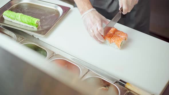 Male Cook Cutting Prepared Philadelphia Sushi Roll on Board in Kitchen Close Up
