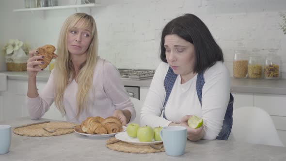 Upset Plump Caucasian Woman Chewing Apple, Her Slim Friend Giving Her Plate with Tasty Croissants