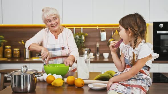 Granny Putting Flour and Egg Into Bowl Mixing It By Whisk Talking to Little Granddaughter Who