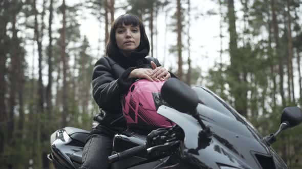 Beautiful Young Caucasian Woman Sitting on Black Motorbike with Pink Helmet and Looking at Camera