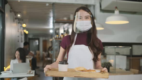 Asian waitress with face protective mask and face shield serving coffee and bread in coffee shop