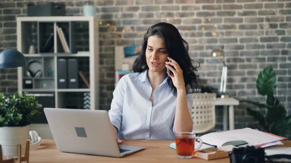 Smiling Girl Talking on Cellphone and Working with Computer in Modern Office