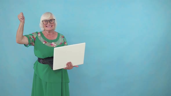Joyful Positive Elderly Woman with a Laptop in Her Hands Learned About the Win on a Blue Background