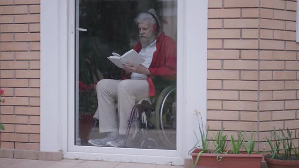 Wide Shot of Happy Engrossed Disabled Senior Man Reading Book Listening to Music in Headphones