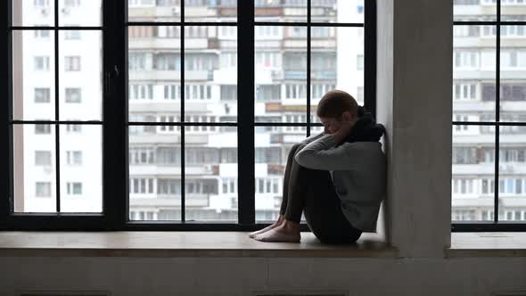 A sickly-looking young woman is sad on the windowsill of a city apartment