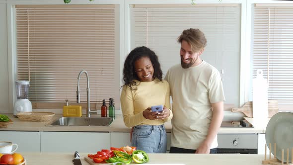 Smiling Heterosexual Diverse Couple Looking at Mobile Phone in Apartment Kitchen