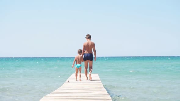 Children Dressed in Palavatelnye Shorts Go To the Azure Sea on a Wooden Pier Against the Blue Sky on