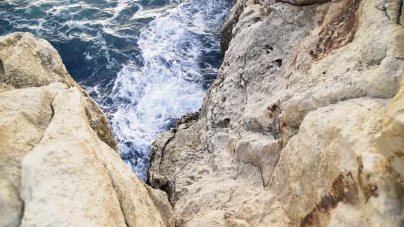 Looking down at blue ocean sea water waves crashing on steep rocky tan cliffs, Malta, zoom out