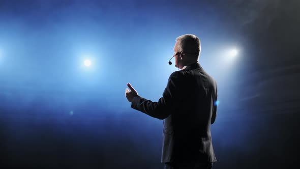 Rear View of Men Speaking Through a Microphone in Dark Conference Hall. Man Talks Into Microphones