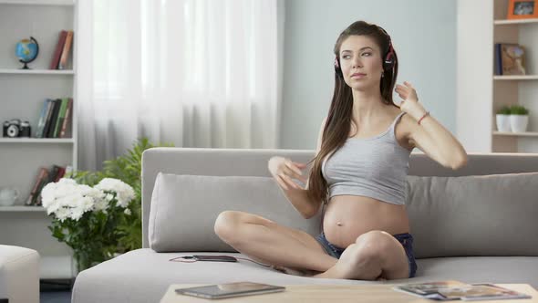 Young Pregnant Woman Sitting on Couch in Headphones, Doing Stretching Exercises