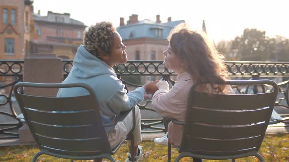 Back View of Diverse Lesbian Couple Sitting on Folding Chairs Outdoors Holding Hands and Kissing