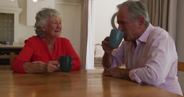 Smiling caucasian senior couple sitting at table in dining room, talking and drinking cups of coffee