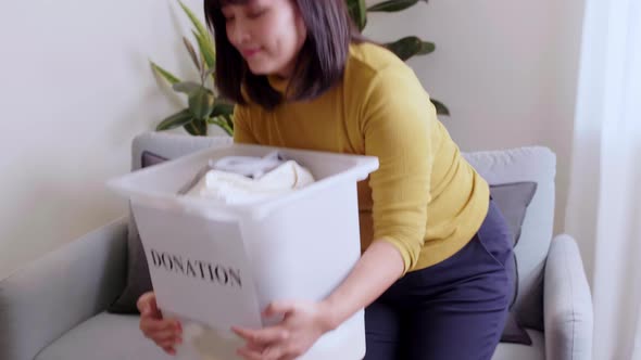 Young Asian woman looking at camera holding box with clothes for donation.