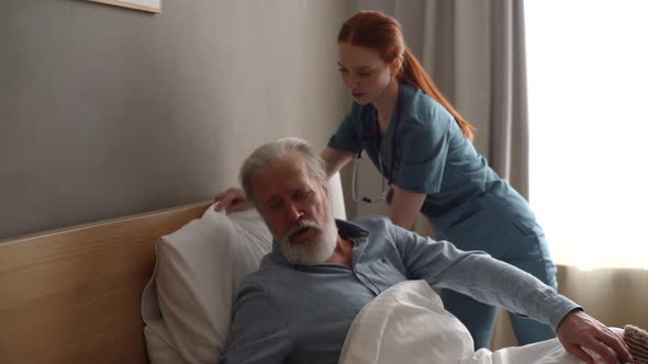 Caring Young Female Home Caregiver Helping Senior Adult Male Patient to Lying on Bed in Bedroom