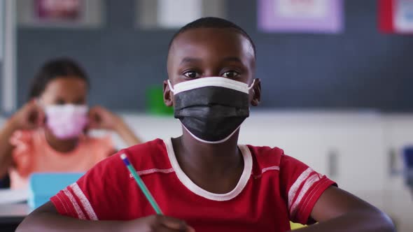 Portrait of african american schoolboy wearing face mask, sitting in classroom looking at camera