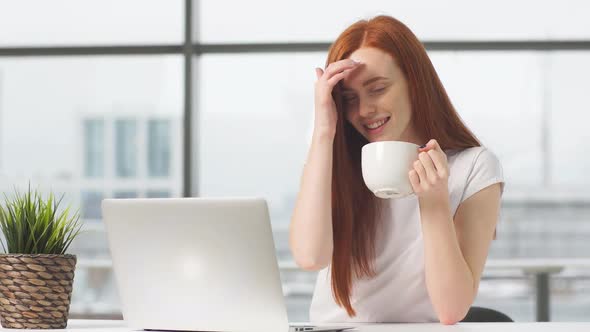 Joyful Redhead Girl Sitting By Table with Laptop and Looking in Camera