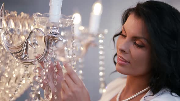 a Brunette in a White Dress and with Pearls Around Neck Touches Crystal Chandelier Pendants